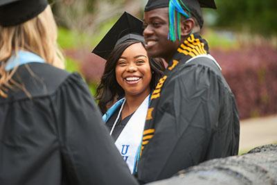 UWF graduates smile while chatting on the Cannon Green as they wear cap and gown.